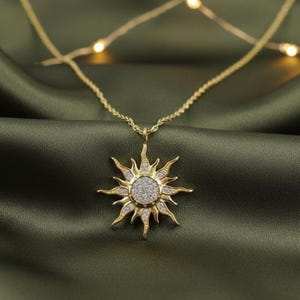 May include: Gold-toned necklace featuring a sunburst pendant. The pendant has a central circle encrusted with small, sparkling stones, surrounded by pointed rays. The chain is delicate, and the background is a dark green fabric.