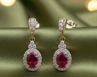 May include: A pair of gold-toned drop earrings with oval, deep red gemstones. Each earring is surrounded by a halo of small, clear stones and features a decorative top section with additional clear stones. The earrings are displayed on a green fabric.