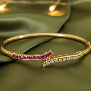 Może przedstawiać: Złota bransoletka typu bangle z zawiasowym designem. Jedna strona ozdobiona jest rzędem małych, żywych czerwonych kamieni szlachetnych, a druga rzędem przezroczystych, błyszczących białych kamieni. Bransoletka spoczywa na zielonej satynowej powierzchni.