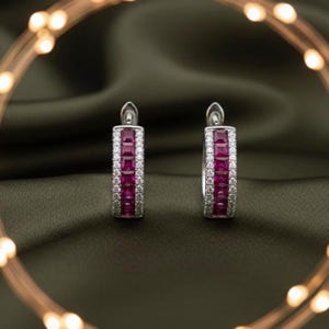 May include: A pair of silver hoop earrings with a rectangular design. The earrings feature a row of square, deep red gemstones in the center, framed by smaller, clear gemstones. The earrings are displayed on a dark green fabric.