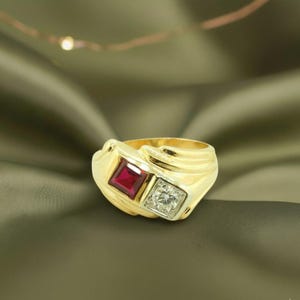 Op de afbeelding: Een gouden ring met een vierkante rode edelsteen en een vierkante heldere edelsteen. De ring heeft een dikke band met een textuurontwerp. De edelstenen staan naast elkaar, wat een gedurfde en elegante uitstraling creëert.