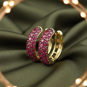 Puede incluir: Un par de pendientes de aro dorados incrustados con numerosas piedras preciosas pequeñas, redondas y de color rubí. Los pendientes se colocan sobre un fondo verde oscuro y sedoso, con un borde de luz borroso y cálido.