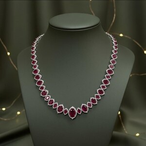 Könnte beinhalten: Halskette mit oval geschliffenen Rubinen und Diamanten, präsentiert auf einer Büste. Die Rubine sind tiefrot und von Diamanten umgeben. Die Halskette ist in einer Silber- oder Weißgoldeinfassung gefasst. Ein luxuriöses Schmuckstück.