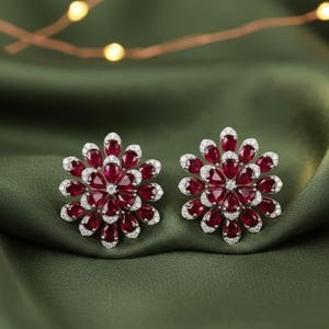 May include: A pair of floral-shaped stud earrings. Each earring features a cluster of deep red, teardrop-shaped gemstones surrounded by smaller, clear gemstones. The earrings are set against a dark green, silky background.