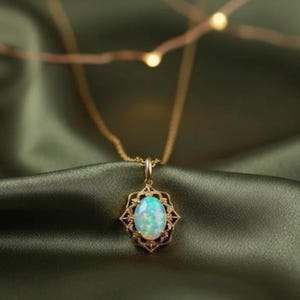 May include: A gold-colored necklace with an oval opal pendant. The opal has a blue-green hue and is set in a decorative, openwork frame. The necklace is displayed on a dark green, satin-like fabric.