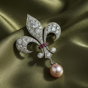 Op de afbeelding: Zilverkleurige broche in de vorm van een lelie, bezet met talrijke heldere edelstenen. Een kleine ring van rode edelstenen bevindt zich in het midden. Een hangende, ronde, roze parel hangt aan de onderkant van de broche. Op een groene achtergrond.