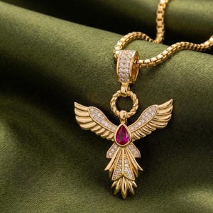 May include: A gold-toned pendant necklace featuring a stylized bird or angel wing design. The piece is adorned with small clear pavé stones and a central pear-shaped red gemstone. It hangs from a matching gold-toned box chain link necklace.