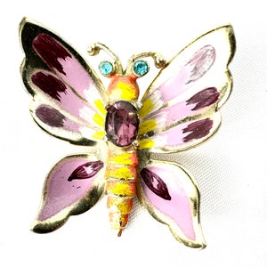May include: A butterfly brooch with a gold-tone metal frame. The wings are painted in shades of pink, white, and burgundy, with a central purple gemstone. The body is yellow and orange, and the antennae have small blue accents.