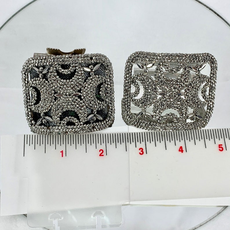 May include: Two silver metal decorative boxes with intricate designs. The boxes are about 2 inches wide and 1.5 inches tall. They are sitting on a white surface with a ruler for scale.