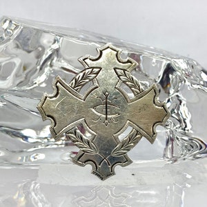 May include: A silver cross pendant with a decorative design of leaves and a monogram in the center.