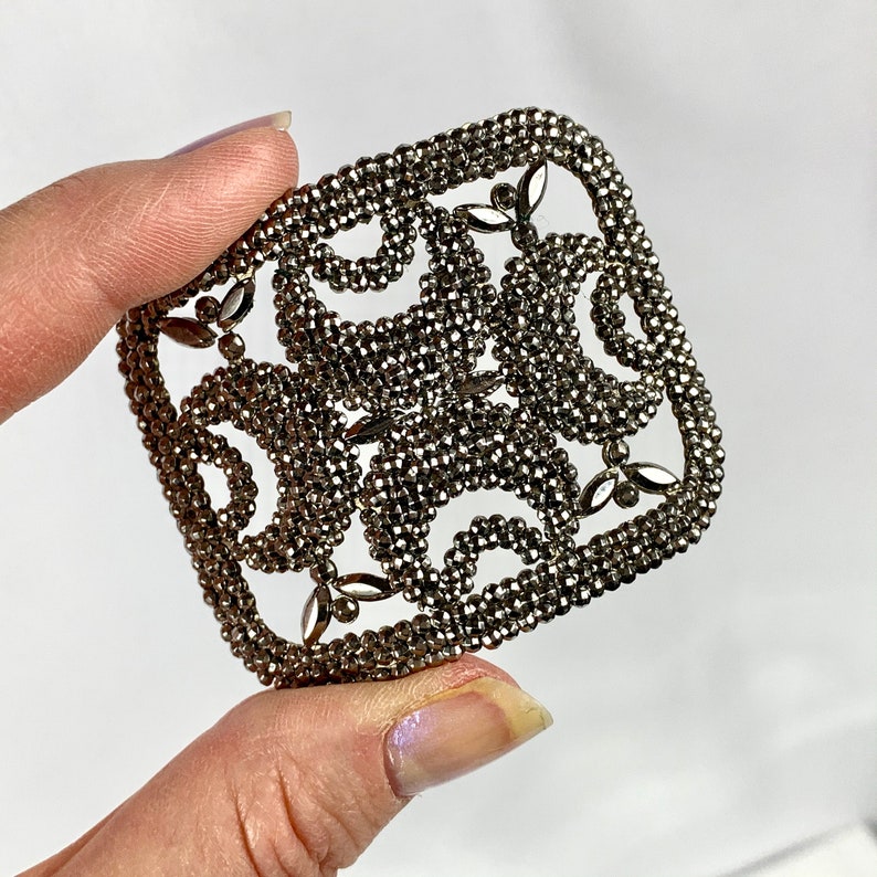 May include: A silver metal brooch with a geometric design. The brooch is made of small beads that are arranged in a pattern of circles and squares.