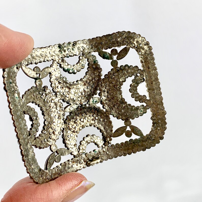 May include: A silver metal decorative buckle with a detailed, intricate design featuring a repeating pattern of circles and swirls.