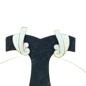 May include: A pair of silver earrings with a white enamel design. The earrings are shaped like a stylized leaf with a swirl at the bottom.