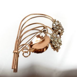 May include: A vintage gold-toned brooch featuring a stylized floral design with clear rhinestones. The brooch has a delicate, intricate design and is a beautiful example of mid-century jewelry.