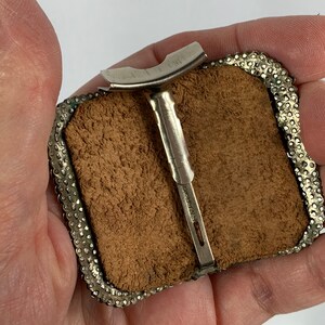 May include: A silver-toned belt buckle with a leather backing and a decorative border. The buckle has a metal clasp and a small metal loop on the back.