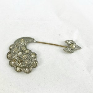 May include: A vintage silver-toned brooch with clear rhinestones. The brooch features a crescent-shaped design with a floral edge and a straight pin with an arrow-shaped end. The brooch is set against a white background.