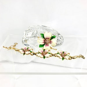 May include: A gold bracelet with white and pink enamel flowers. The bracelet has a delicate chain and is accented with green leaves. The flowers have a sparkling center.