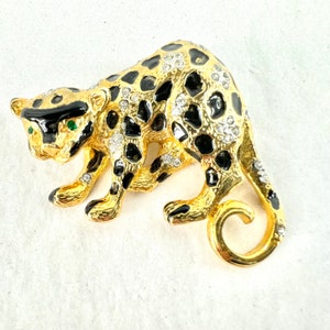 May include: A gold-tone brooch in the shape of a leopard with black spots and green eyes. The leopard is decorated with rhinestones.