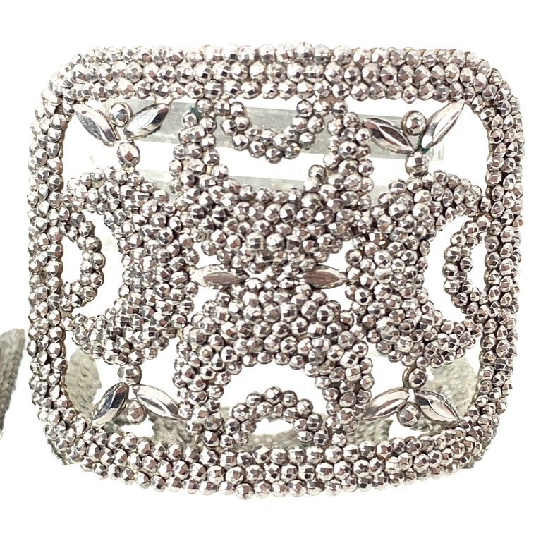 May include: A silver square-shaped bracelet with a delicate, intricate design made of small beads. The bracelet has a geometric pattern with open spaces.