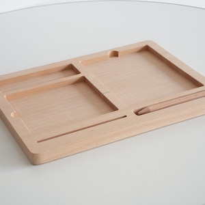 May include: A light-colored wooden desk organizer with multiple compartments and a slot for a pencil. The organizer has a rectangular shape with a smooth, natural wood grain finish. The compartments are various sizes and shapes.