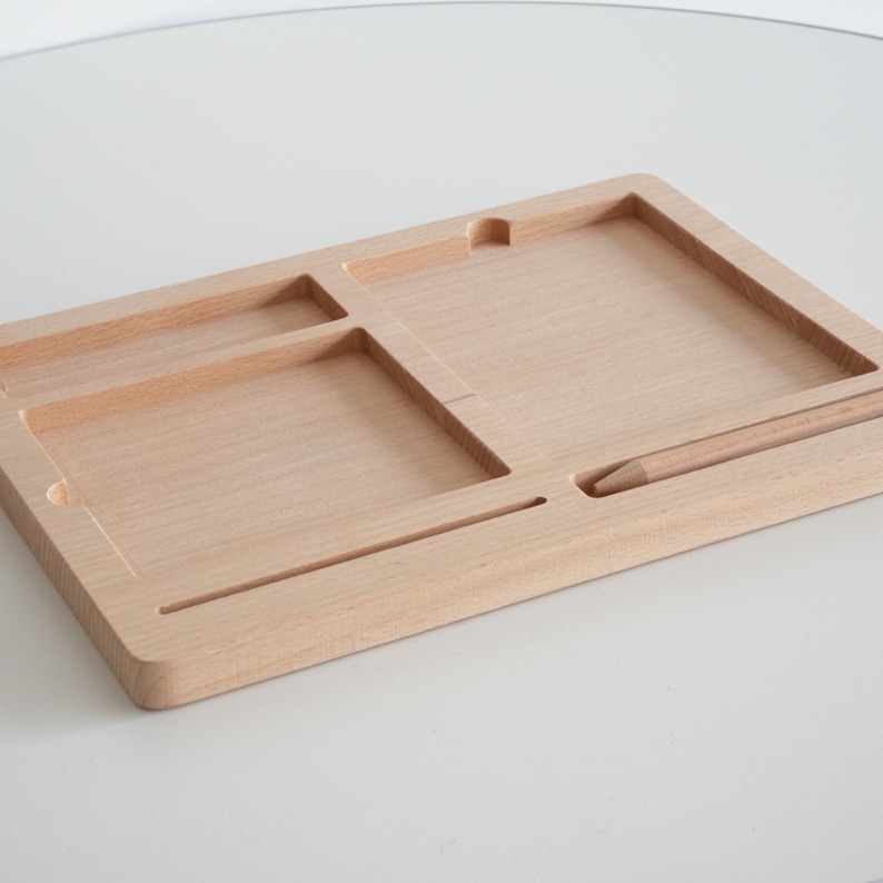 May include: A light-colored wooden desk organizer with multiple compartments and a slot for a pencil. The organizer has a rectangular shape with a smooth, natural wood grain finish. The compartments are various sizes and shapes.
