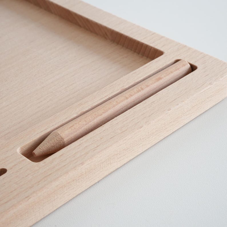 May include: A light-colored wooden desk organizer with a recessed area and a slot holding a pencil. The organizer has a rectangular shape with a smooth, natural wood grain finish. The pencil is also made of wood.