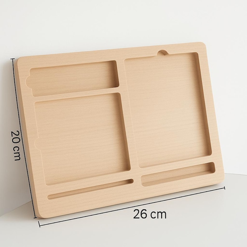 May include: A light-colored wooden lap desk with multiple compartments. The desk measures 26 cm long and 20 cm wide. It features a handle-like cutout and various rectangular and curved slots for organizing items. The desk is designed for use on a lap or flat surface.