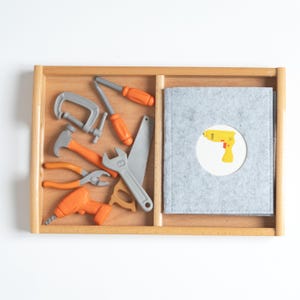 May include: A wooden tray containing a set of plastic toy tools including a grey C-clamp, an orange screwdriver, a hammer, pliers, a drill, and a grey saw, alongside a grey felt folder featuring a circular cutout with a yellow drill graphic.