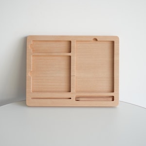 May include: A light-colored wooden desk organizer with multiple compartments. The organizer has a rectangular shape with a variety of slots and sections for storing items. The wood grain is visible, and the design is simple and functional.