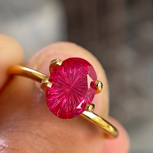 Könnte beinhalten: Ein Goldring mit einem ovalen, leuchtend pinkfarbenen Edelstein, der ein ausgeprägtes, strahlenförmiges Facettenschliffmuster aufweist. Der Stein wird von vier kleinen Goldkrappen auf einer dünnen, polierten Goldschiene gehalten.