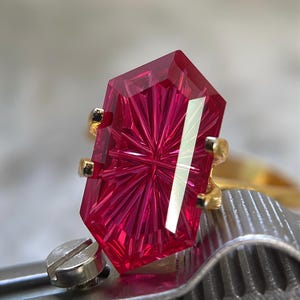 Puede incluir: Una gema de corte hexagonal en color rosa intenso y vibrante montada en un anillo de oro. La piedra presenta un complejo patrón de facetas en forma de estrella que irradia desde el centro. El anillo es sostenido por una herramienta metálica.