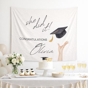 May include: White graduation party backdrop with the text "she did it!" and "Congratulations Olivia". A black graduation cap with a gold tassel is featured. A cake and cupcakes are on the table.