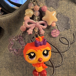 May include: A decorative keychain featuring a small orange bird figurine with large eyes and a purple bow. The charm is attached to a silver metal ring adorned with various beads, including a cream-colored seashell, a star, a purple bow, and a translucent purple star-shaped charm.
