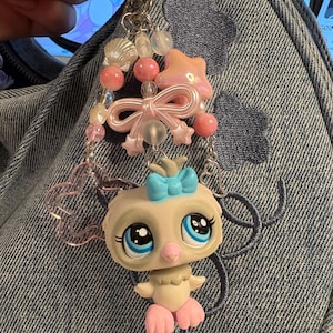 May include: A bag charm featuring a small owl figurine with large blue eyes and a light blue bow on its head. The charm is attached to a silver-toned metal clip and decorated with various pink and clear beads, a pink star, a pink bow, and a seashell.