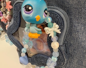 May include: A blue bird-shaped charm with a beaded bracelet, attached to a denim bag with a gold ring. The bracelet features light blue, white, and clear beads, with a white bow and butterfly accents. The charm has a yellow beak and orange feet.