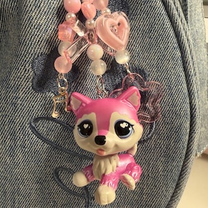 May include: A bag charm featuring a small pink and white plastic animal figurine with large eyes, attached to a silver-toned metal chain. The charm is decorated with various pink and white beads, a translucent pink heart, and a small gold-toned music note charm.