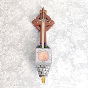 May include: A wooden beer tap handle with a cross design and a sword through the center. The handle has a stone-like base with the text "Dirty Penny Ale" and a copper-colored coin image. The tap is from Olde Brunswick Brewing Co.