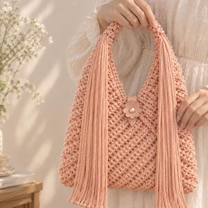 May include: A peach-colored macrame handbag featuring a textured knotted pattern, two long decorative fringe tassels hanging from the handles, and a central leather-like tab closure adorned with a small flower and a gold-toned stud.