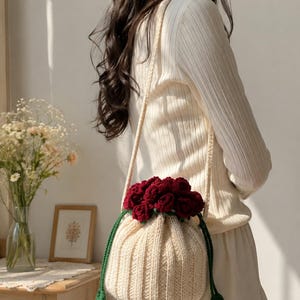 May include: A cream-coloured crocheted bucket bag featuring a drawstring closure adorned with a cluster of deep red crocheted roses and green leaves. The bag has a long, thin crocheted shoulder strap and a ribbed texture throughout the main body.