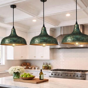 May include: Three pendant lights with a distressed green finish and gold interior hang above a kitchen island. The lights have a bell shape and are suspended from black cords. The kitchen features white cabinets and a stainless steel range.