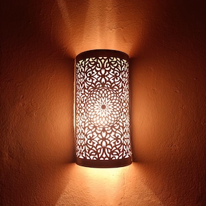 Peut inclure: Applique murale cylindrique avec un motif décoratif en métal ajouré. L'applique est éclairée de l'intérieur, projetant des motifs complexes de lumière et d'ombre sur le mur environnant. Le mur est de couleur chaude et terreuse.