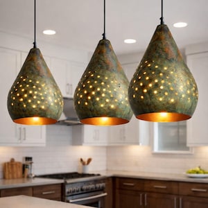 May include: Three teardrop-shaped pendant lights with a mottled green and brown finish hang in a kitchen. Each light has numerous small holes that emit a warm glow. The interior of each light is a bright gold color.