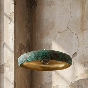 May include: A pendant light fixture with a bronze-colored exterior and a gold-colored interior. The exterior has a green patina. The light hangs from a black cord. The fixture is suspended in front of a textured, aged wall.