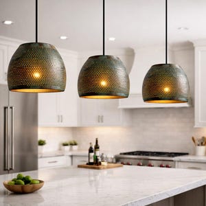 May include: Three pendant lights with a perforated metal shade in a distressed teal finish hang above a kitchen island. The interior of each shade has a gold finish, and the lights are suspended from black cords. The kitchen features white cabinets and a marble countertop.
