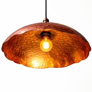 May include: A copper pendant light fixture with a hammered texture. The light has a scalloped edge and a warm, glowing bulb. The fixture hangs from a black cord and is set against a white background.