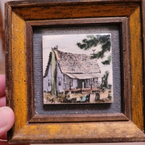 May include: A framed miniature painting of a weathered house with a porch, set against a backdrop of trees and sky. The artwork is in a wooden frame with a gold finish, and the painting is signed in the lower left corner.