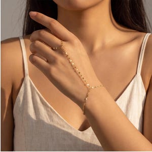 Könnte beinhalten: Eine goldfarbene Handkette mit einem zarten Design aus flachen Gliedern. Das Schmuckstück besteht aus einem Ringelement, das durch eine schmale Kette mit einem Armband verbunden ist und eine durchgehende Struktur über dem Handrücken bildet.