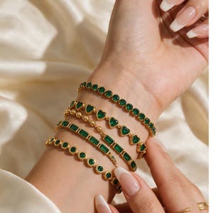 Può includere: Un set di cinque bracciali color oro con pietre verdi. I design includono una catena con pietre rotonde, una catena con pietre a forma di cuore, una catena con piccole perline dorate, una catena con pietre rettangolari e una catena con pietre rotonde incastonate in oro.
