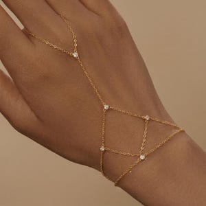May include: A gold-toned hand chain featuring a delicate link design that connects a ring component to a wrist bracelet. The jewelry piece is accented with five small, clear, round-cut stones set in metal bezels at the junction points of the chain.