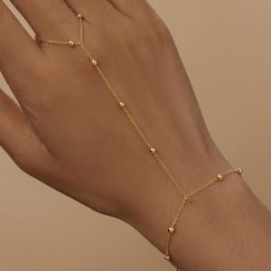 Puede incluir: Una cadena de mano en tono dorado que presenta una delicada cadena de eslabones adornada con pequeñas cuentas esféricas espaciadas uniformemente. La pieza de joyería conecta un anillo en el dedo con una sección de pulsera que rodea la muñeca.